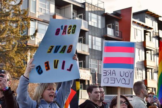 Two people holding signs: One with the transgender flag with "You are loved" and another with a sign reading "GSA SAVES LIVES"